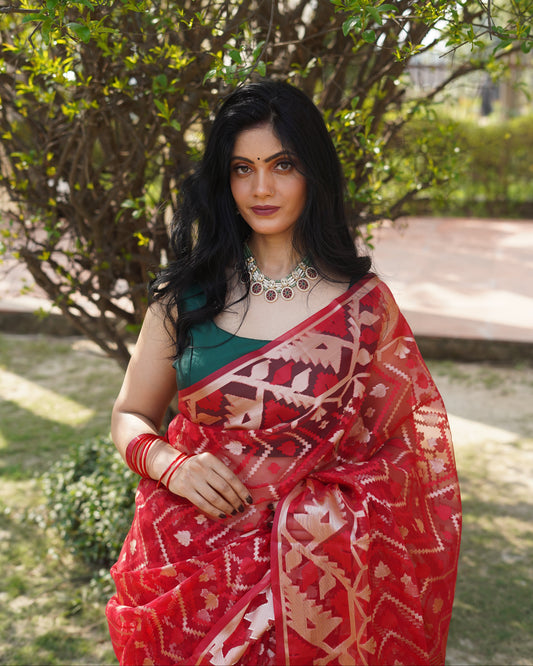 Woman in a red and white patterned saree standing outdoors with greenery in the background