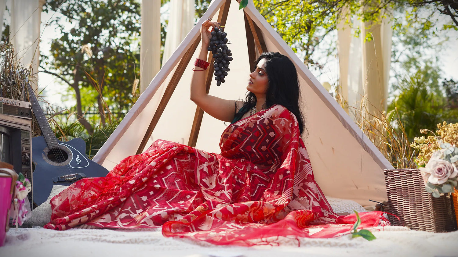 Woman in a red saree sitting inside a teepee with a guitar and basket in the background