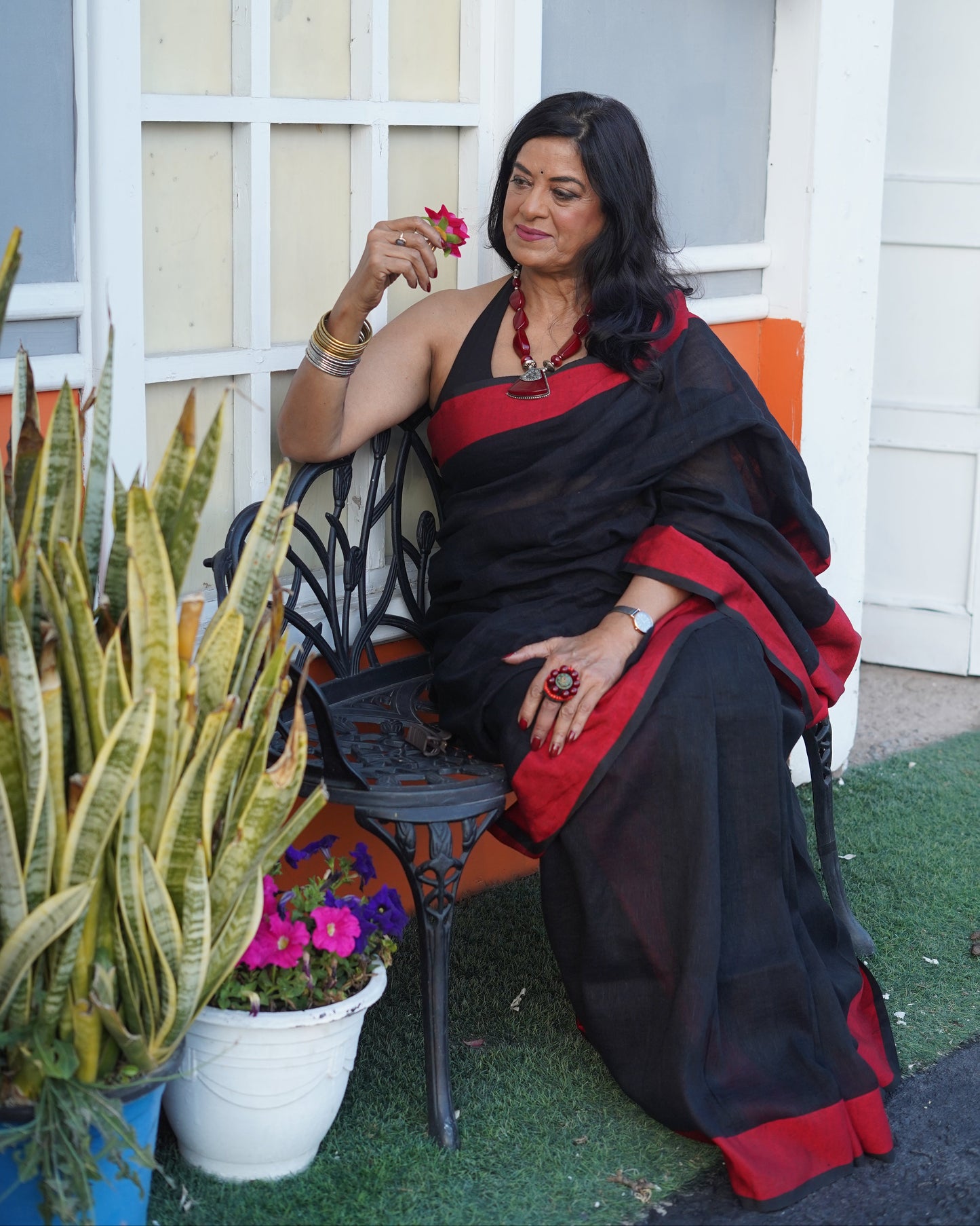 Woman in a black and red saree standing in front of a white door with plants and a colorful rug.