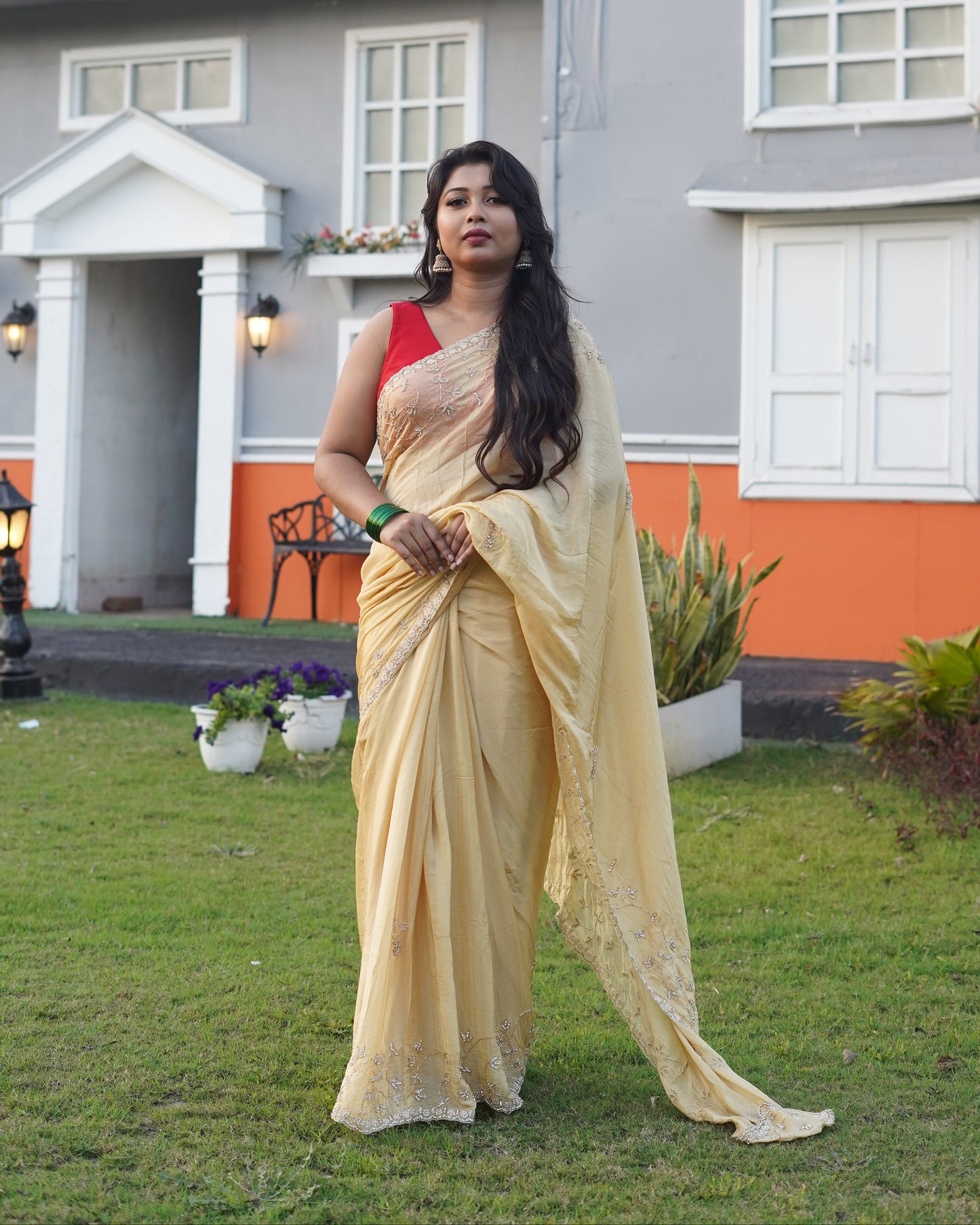 Woman in a yellow saree standing in front of a house with a garden.