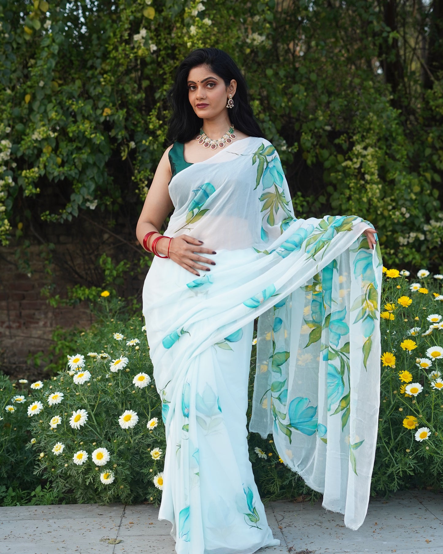 Woman in a white saree with blue floral patterns standing outdoors with greenery and flowers in the background