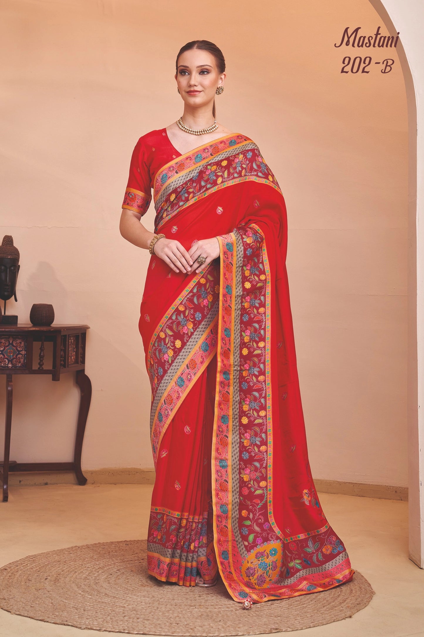 Woman wearing a red saree with colorful patterns, standing against a beige wall.