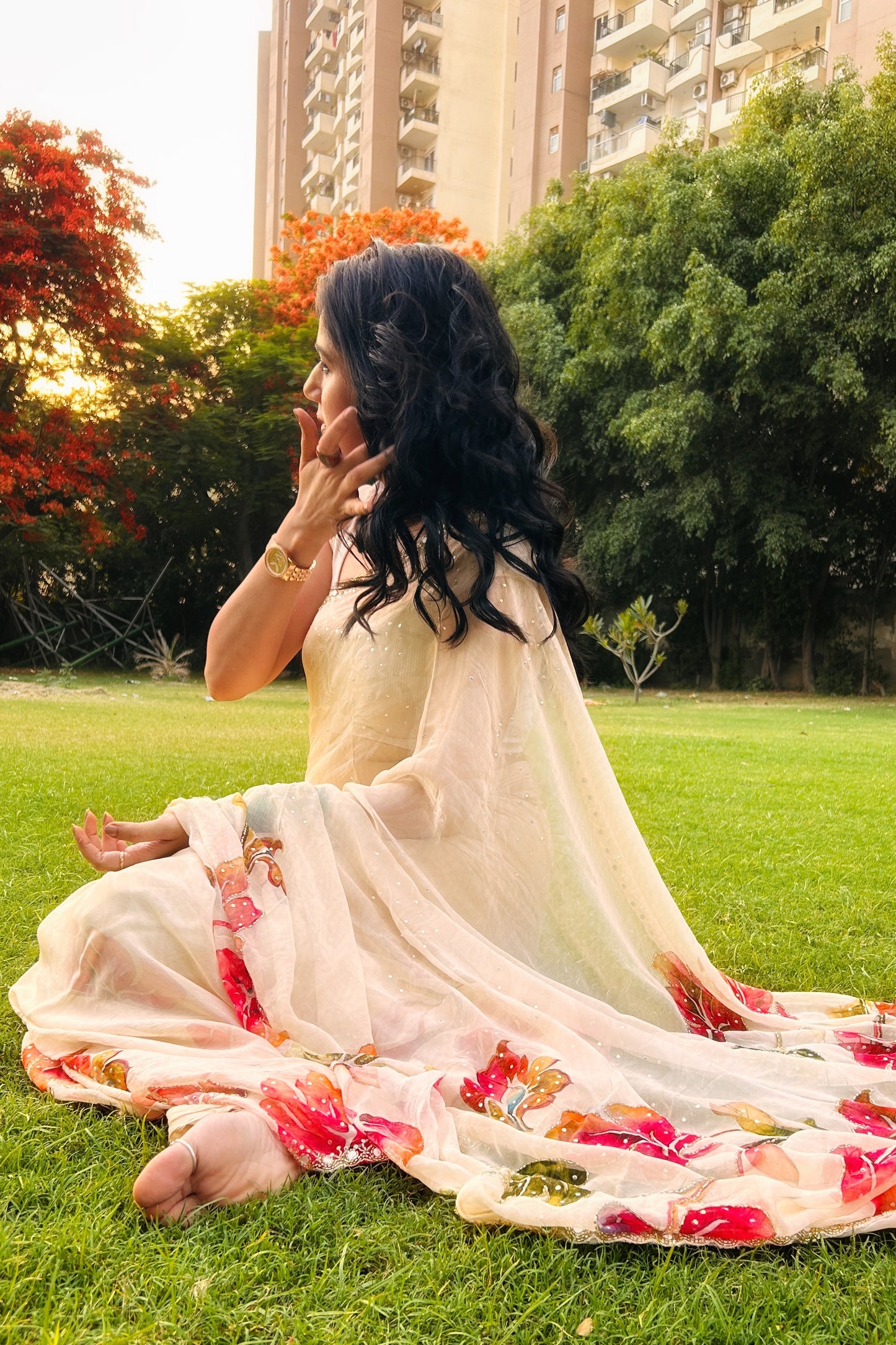 Woman in a floral saree sitting on grass with trees and buildings in the background