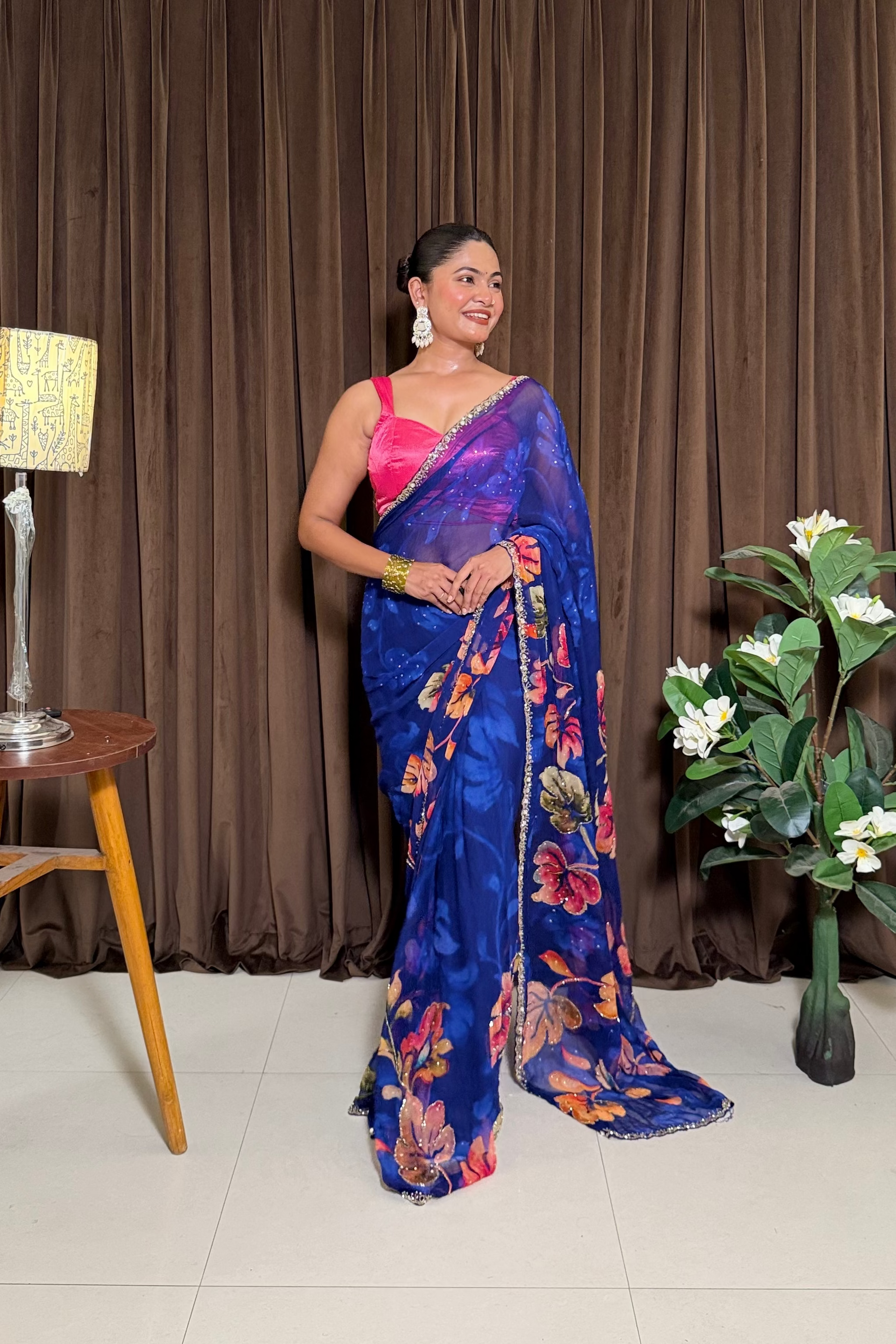 Woman in a blue saree standing in front of a brown curtain with a lamp and plant on either side.