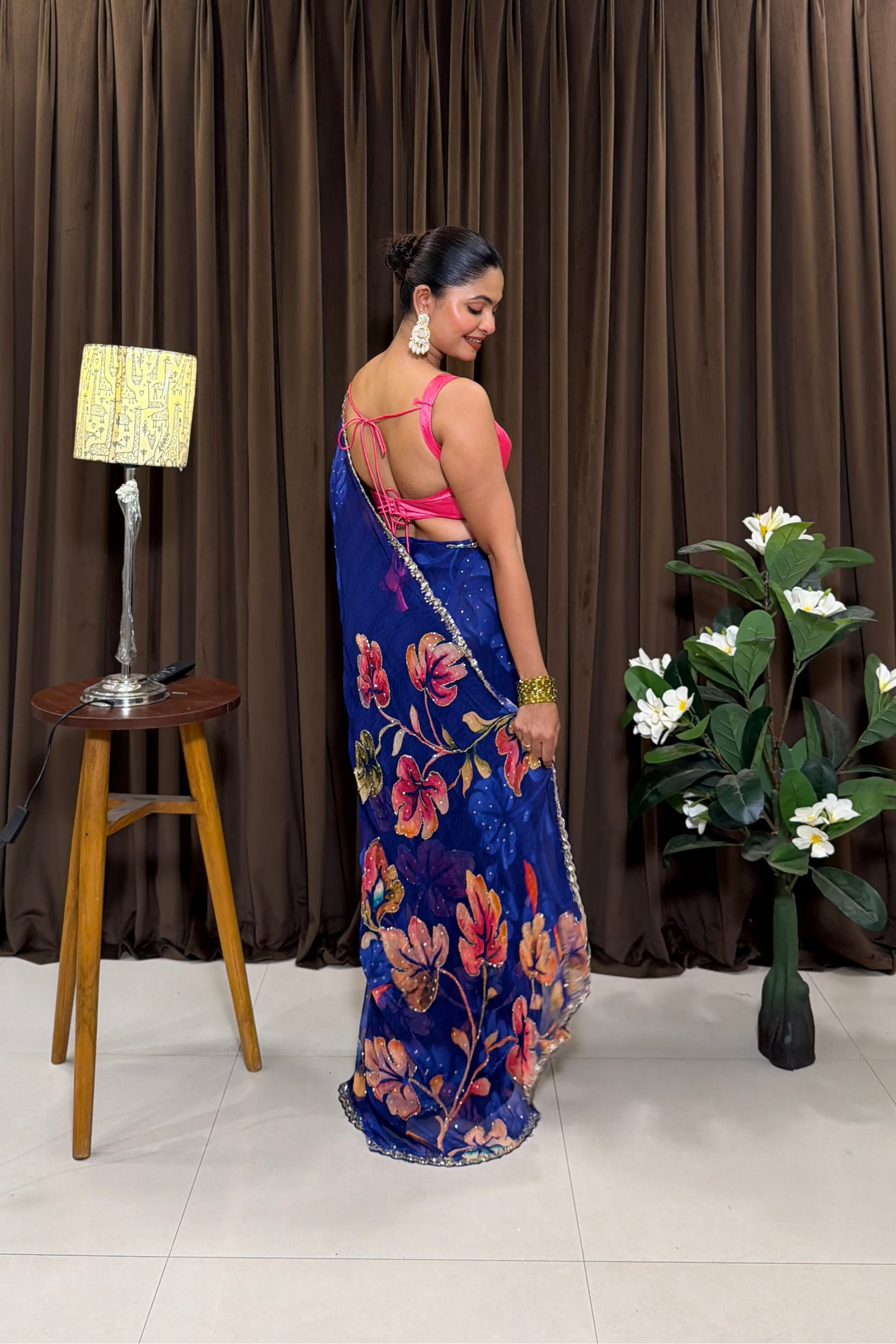Woman in a blue floral saree standing in front of a brown curtain with a lamp and plant on a stool.