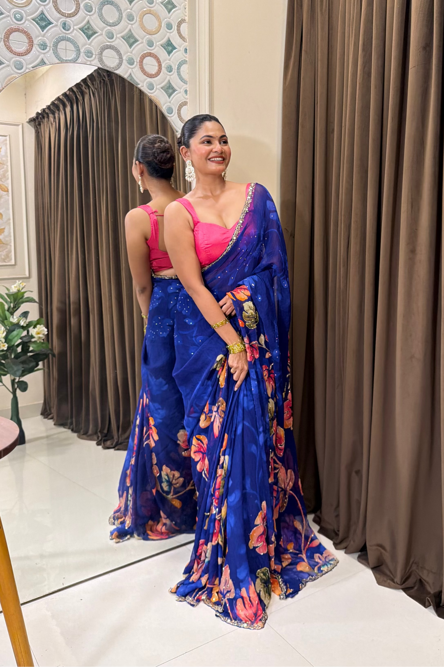Woman in a blue saree with floral patterns standing in front of a mirror.