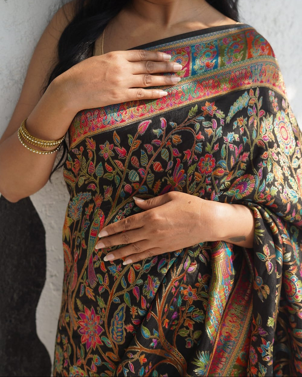 Woman wearing a black saree with colorful floral patterns against a gray wall.