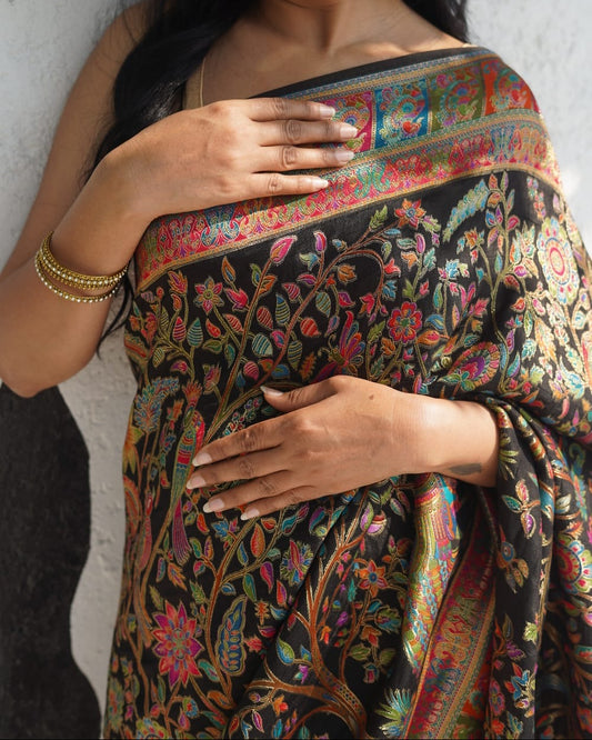 Woman wearing a black saree with colorful floral patterns against a gray wall.